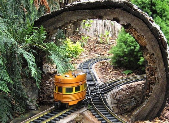 Garden area with a miniature train and tracks going under a tree stump bridge.