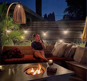 Man sitting at night by a fire pit on patio furniture.
