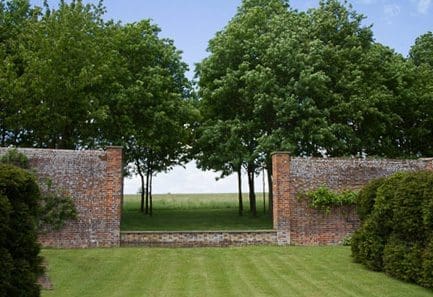 Brick wall with a large opening leading to a large field with large trees.