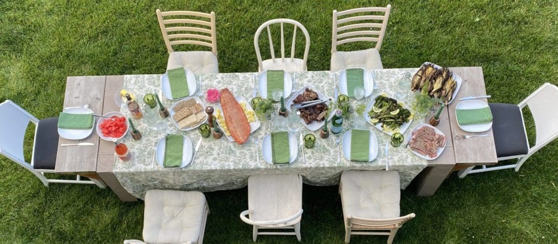 An outdoor wooden dining table set with white plates and green napkins with various dinner foods served.